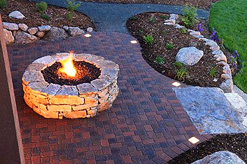 Stone fire pit with flames on a curved paver terrace surrounded by planting, built by Moss Landscaping